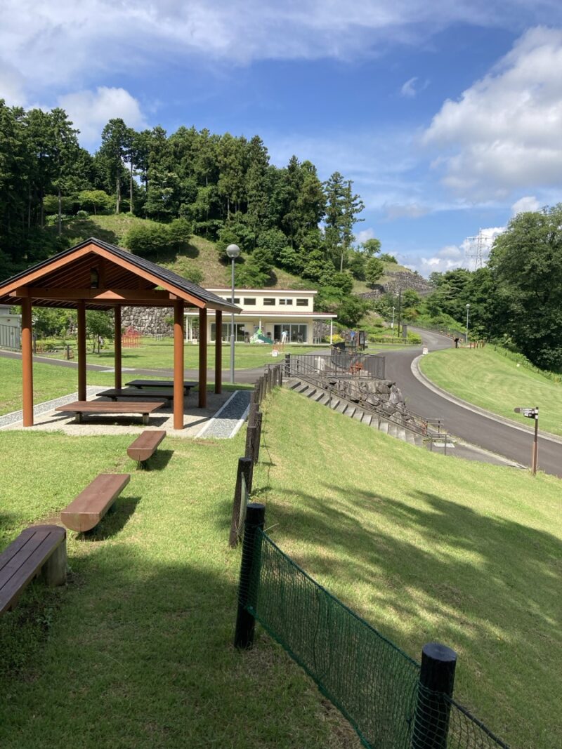公園の芝生広場にある屋根付き休憩所とベンチ 奥に建物と坂道