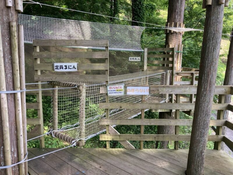 森の中にある木製ツリーハウス遊具 ネットと木の板の通路 定員3人の看板