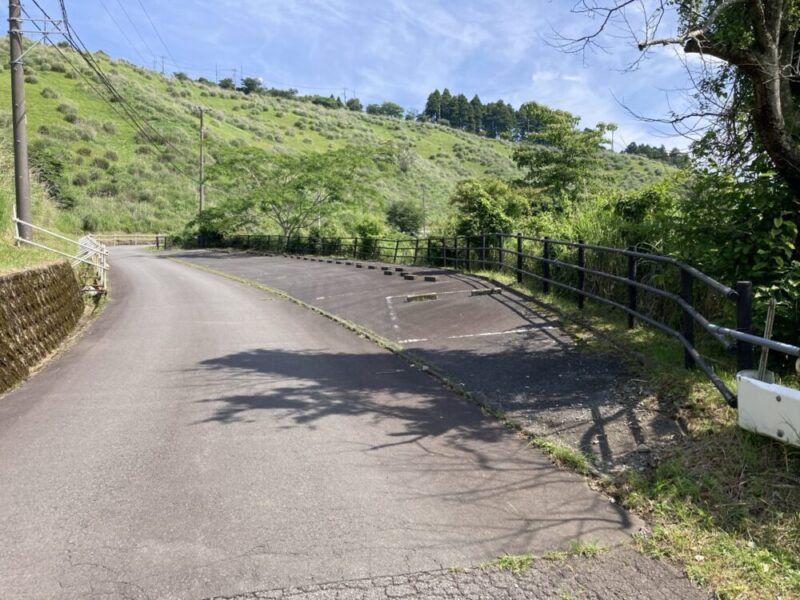 山道をカーブした先に、 grassy な丘が広がり、空は晴れている