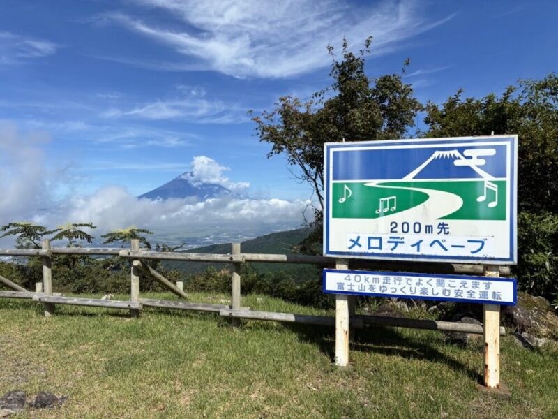 雲をまとった富士山を背景に 道路と音符の絵が描かれたメロディペープの案内標識 200m先で40km走行時に富士山を楽しみ安全運転を促す表示