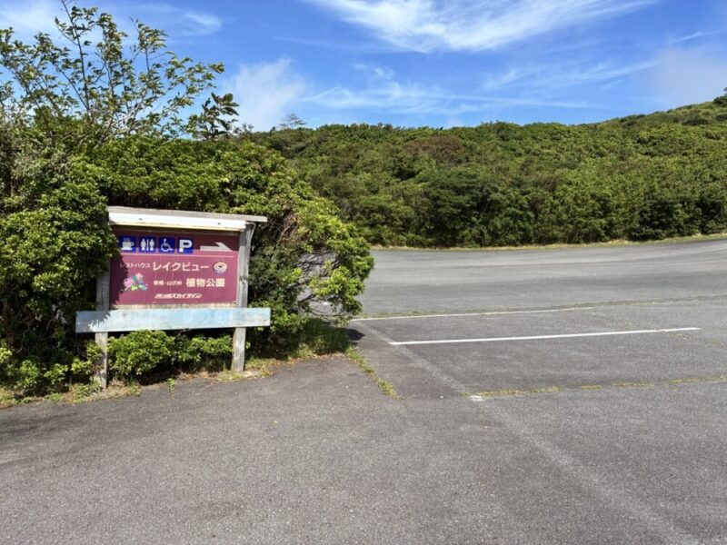 青い空と緑の山々が見える駐車場に建つ案内板 トイレ 車椅子 駐車場 レストランハウスレイクビュー 植物公園の表示と右を指す矢印がある