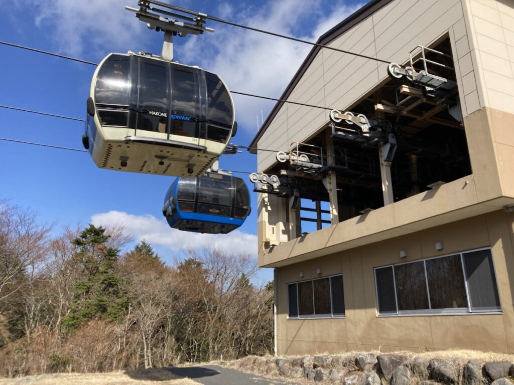 箱根ロープウェイの駅舎に近づくクリーム色と青色のゴンドラと青空 枯れ木の風景