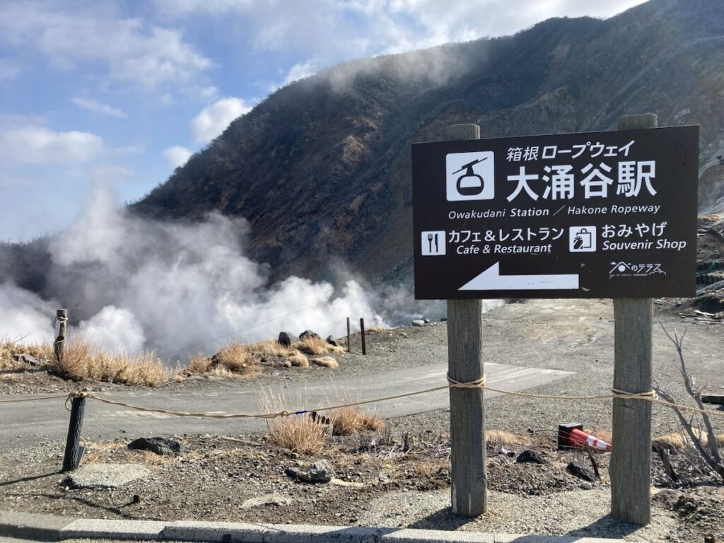 箱根大涌谷駅を示す案内看板と噴煙を上げる山肌