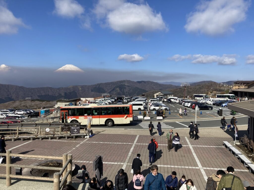 青空の下、雪化粧した富士山を遠景に、バスや車で賑わう展望台を多くの人が歩いている
