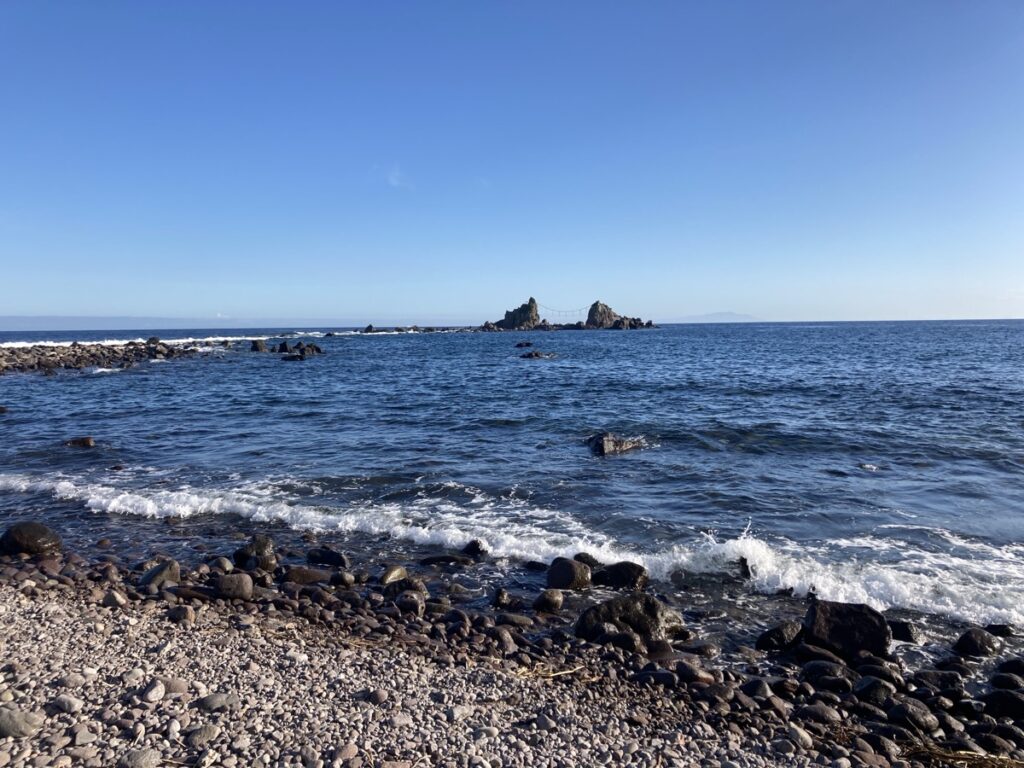 小石の海岸に波が打ち寄せ沖に二つの岩を繋ぐ橋が見える青い海