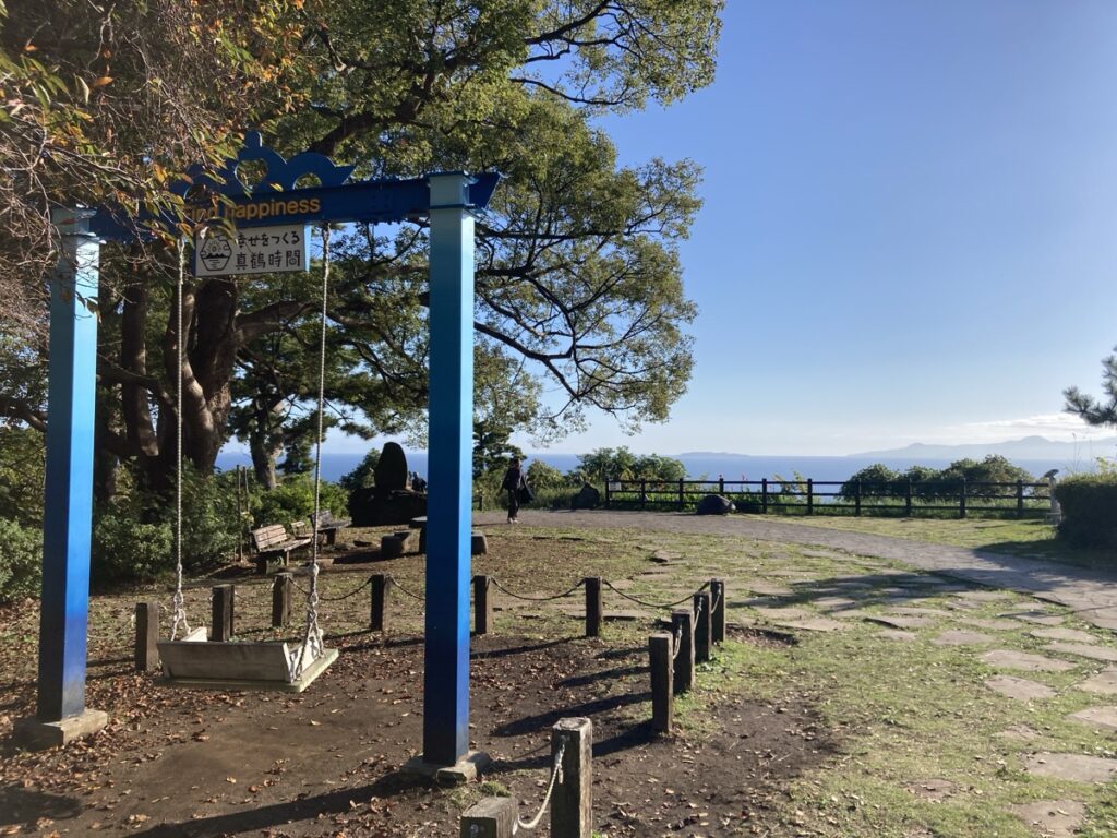 「Happiness幸せをつくる夢語時間」の看板と青い支柱のブランコがある広い海と青空が見える高台の公園