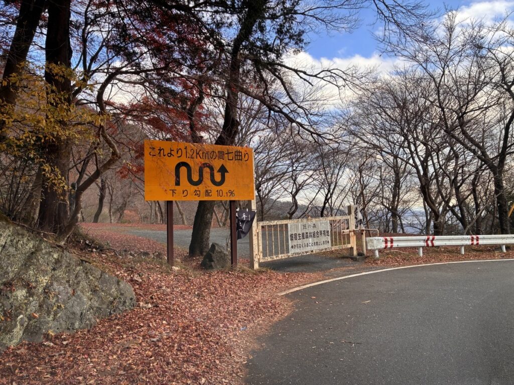 紅葉と落ち葉が積もる秋の山道で1.2キロ続く七曲りの急な下り坂を示すオレンジ色の標識があるカーブ