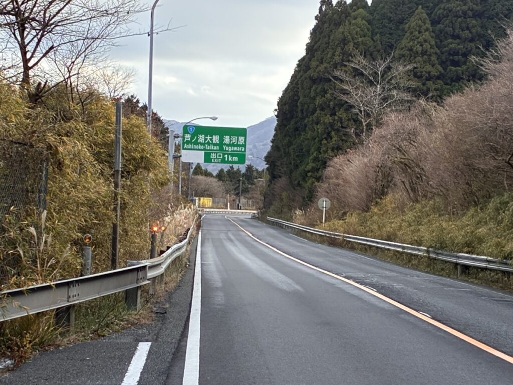 冬の日本の道路沿いにある、芦ノ湖大観と湯河原方面の出口まで1kmを示す緑色の案内標識