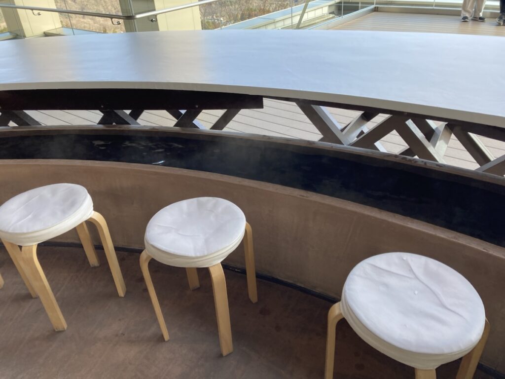 白い座布団付きの丸椅子が3脚並び奥に湯気立つ足湯がある屋外テラス